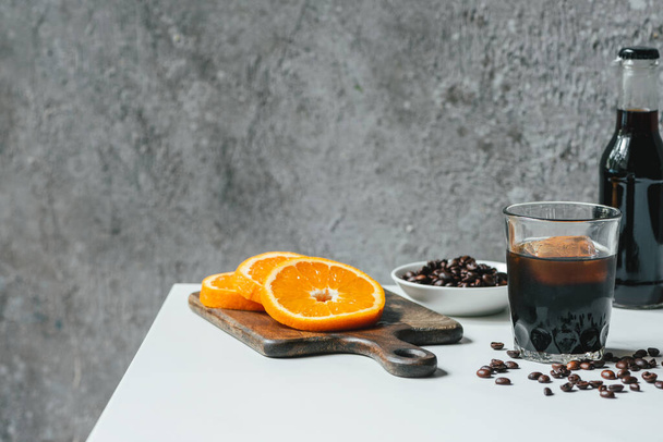 Buzlu soğuk demlenmiş kahve ve portakal dilimlerinin yanında şişe ve beyaz masada kahve çekirdekleri. - Fotoğraf, Görsel