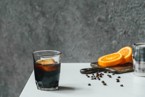 selective focus of cold brew coffee with ice in glass near orange slices on chopping board and coffee beans on white table - Фото, изображение