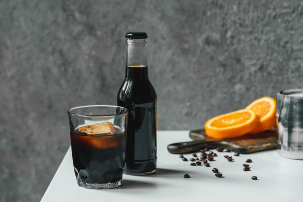 selective focus of cold brew coffee with ice in glass and bottle near orange slices on chopping board and coffee beans on white table - Valokuva, kuva