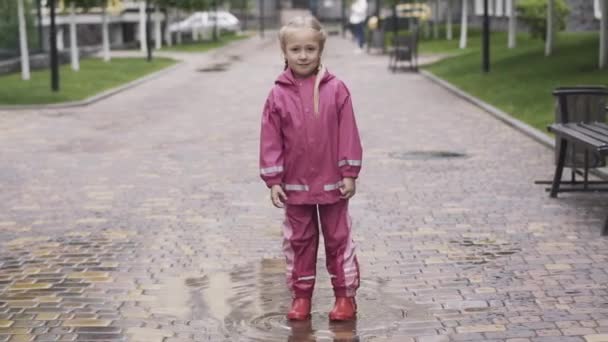 Portrait of cute little girl in pink waterproof costume and rubber boots standing in puddle and looking at camera. Pretty blond Caucasian kid posing outdoors on rainy season. Fun, childhood, lifestyle - Video