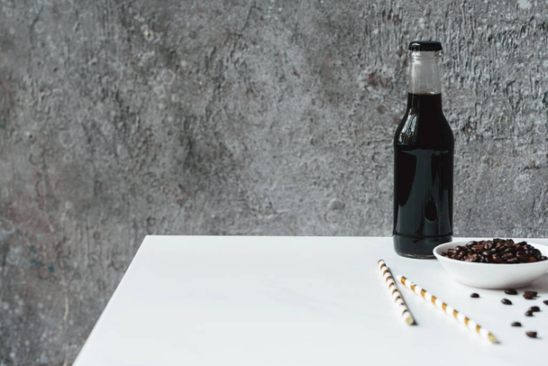 selektivní zaměření studené kávy s ledem v láhvi v blízkosti pitné slámy a kávová zrna na bílém stole - Fotografie, Obrázek