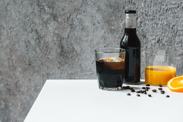 cold brew coffee with ice in glass and bottle near orange juice and coffee beans on white table - Φωτογραφία, εικόνα