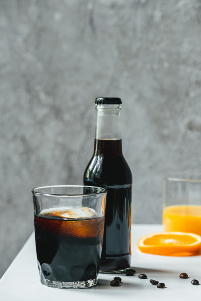 selective focus of cold brew coffee with ice in glass and bottle near orange juice and coffee beans on white table - Foto, afbeelding
