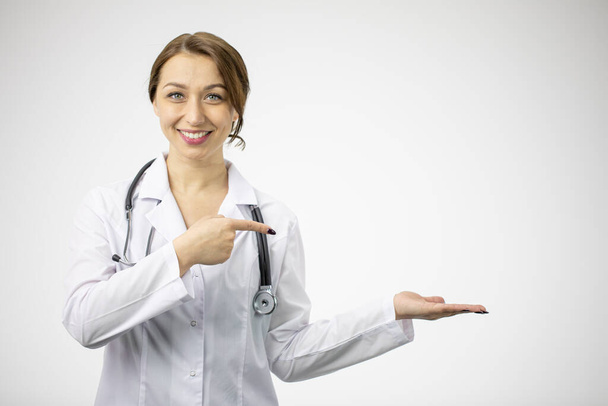 Smiling sexy doctor in medical uniform points with finger to empty open palm - Фото, изображение