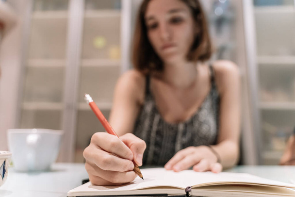 Selektywne skupienie ręki dziewczyny z ołówkiem w notatniku w stole z filiżanką wewnątrz pokoju - Zdjęcie, obraz