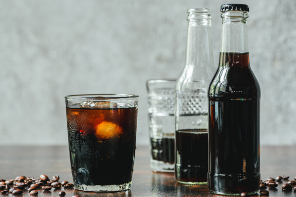 selective focus of cold brew coffee with ice in glass and bottles near coffee beans - Zdjęcie, obraz