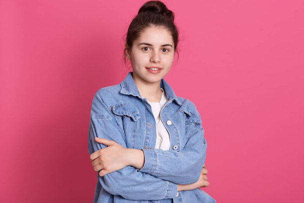 Young girl with pleasant appearance standing against pink wall, dresses denim jacket and white shirt, keeps hands folded, looks at camera, expressing confidence. - 写真・画像