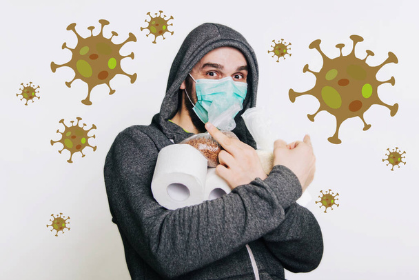 Ein Typ in grauem Sweatshirt auf hellem Hintergrund mit medizinischer Maske hält seinen Mittelfinger und zeigt einen Fac. Ein Mann in Panik kauft Toilettenpapier und Buchweizengrütze, Reis. - Foto, Bild
