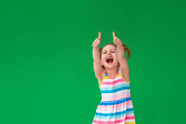 small, mischievous, laughing girl, Caucasian, with light hair on green background. child is having fun and shows his thumbs up, a sign of approval. concept luck and appreciation. Carefree childhood. - Фото, изображение