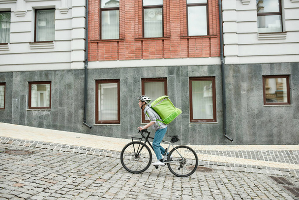 Young delivery woman in helmet with thermo bag or backpack riding a bike along the city, delivering food. Courier, delivery service concept - Фото, изображение