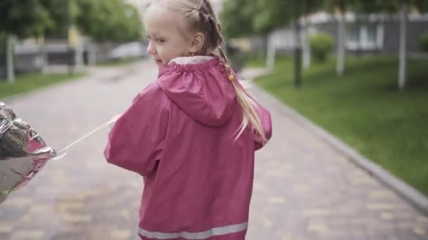 Back view of cheerful blond girl with balloon riding scooter. Portrait of Caucasian kid enjoying weekends outdoors on rainy day. Camera moves down from face to feet. Childhood, leisure. - Materiał filmowy, wideo
