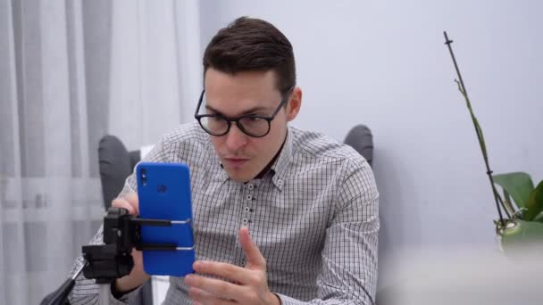 Young attractive man wear plaid shirt, black glasses sit on chair, check social feed on blue phone with tripod, touch screen, watch videos close up. Male goes live, camera ready for online teaching. Inside view. - Footage, Video