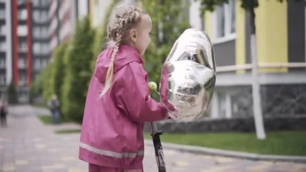 Side view of cheerful girl with pigtails riding scooter. Happy pretty Caucasian child with balloon enjoying resting outdoors. Leisure, joy, relaxation, childhood. - Video