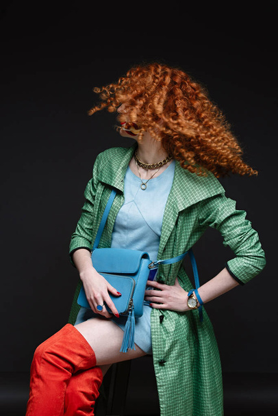  girl with red hair and white skin, bows her head, curly curly hair, lush hairstyle, red lipstick, beautiful makeup, green polto and orange long boots - Foto, imagen