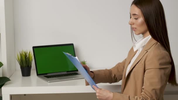 Kaunis brunette valkoihoinen ulkonäkö työskentelee toimistossa pöydässä pitämällä liukupöydällä asiakirjoja ja tarkistaa tiedot kannettavalla tietokoneella, jossa on näyttö vihreällä taustalla - Materiaali, video
