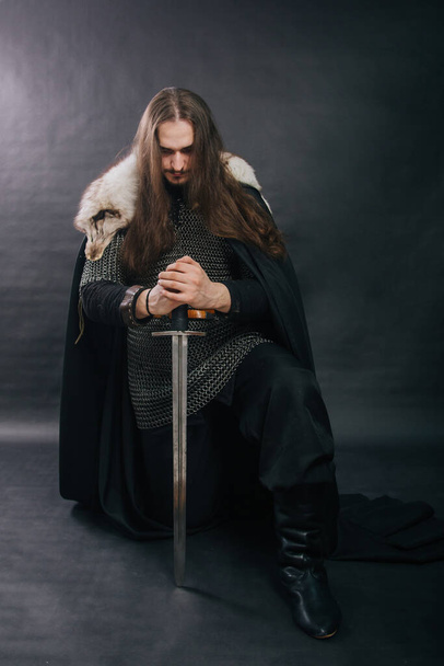 Warrior in armor with a sword. A guy with long hair and a beard, a fox collar. Dressed in chain mail, a black cloak and black pants holds a sword in his hands while sitting on one knee. - Photo, Image