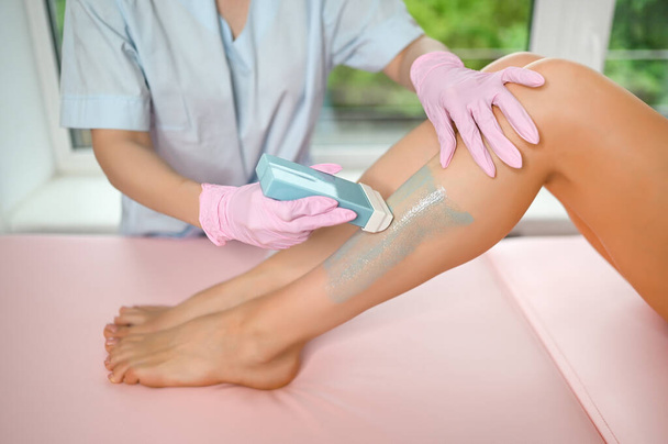 Woman with long tanned perfect legs and smooth skin having wax stripe depilation hair removal procedure on legs in beauty salon. Beautician in blue robe, pink gloves. Body care, epilation spa concept. - Photo, Image