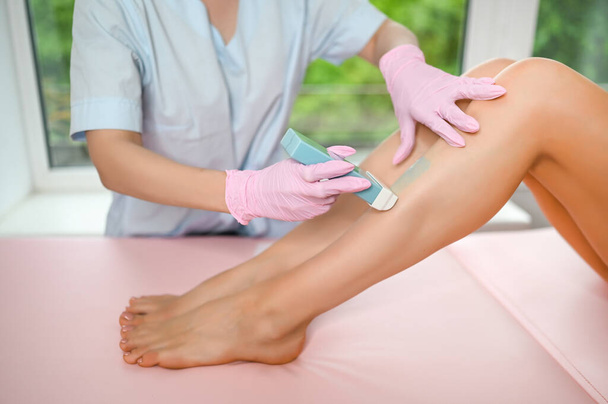 Woman with long tanned perfect legs and smooth skin having wax stripe depilation hair removal procedure on legs in beauty salon. Beautician in blue robe, pink gloves. Body care, epilation spa concept. - Foto, imagen