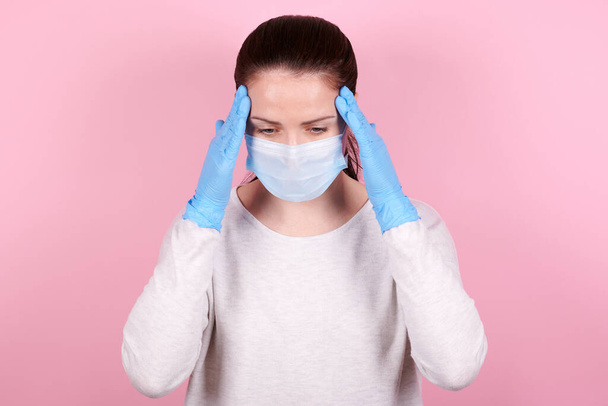 Portrait of a brunette girl with headache in a medical mask and blue rubber gloves. Isolated on pink background. - Foto, imagen
