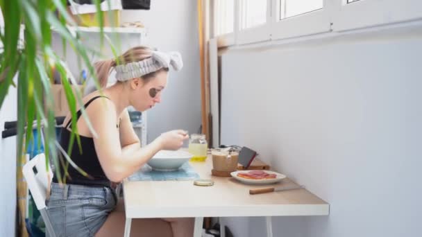 Μια νεαρή ελκυστική ξανθιά γυναίκα με μαύρο μπλουζάκι, τζιν σορτς φορούν κορδέλα μαλλιών, κάθονται στο τραπέζι, τρώνε ζεστό χυλό, βλέπει βίντεο στο smartphone στο λευκό μπαλκόνι στο σπίτι. Πρωινό πρωινό. Πλευρική άποψη. - Πλάνα, βίντεο