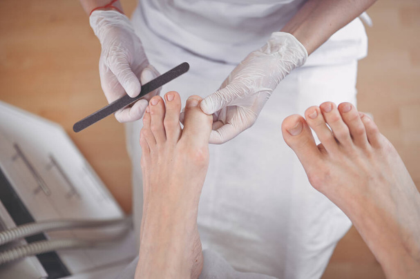 Pedicurist doing professional medical pedicure procedure in beauty salon with nail file. Foot treatment in SPA salon. Podiatry clinic. Beautician doctor hands in white gloves with female legs - Photo, Image
