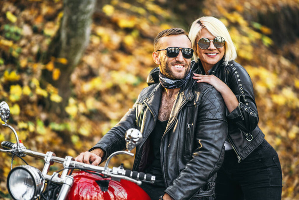 Pretty couple near red motorcycle on the road in the forest with colorful blured background. Relationship concept. - Fotó, kép