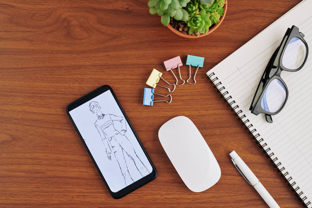 Fashion sketch on screen of smartphone that lies on wooden table next to textbook, glasses and wireless mouse, view from above - 写真・画像