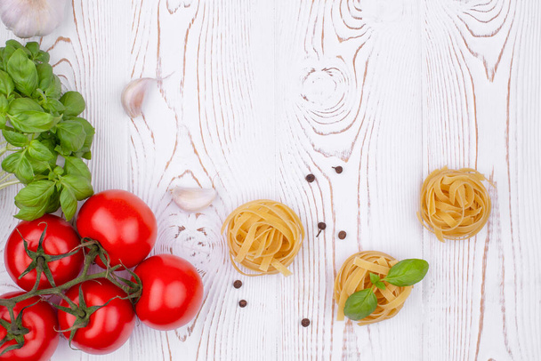 Vista superior pasta de tagliatelle cruda con albahaca fresca, ajo y tomates sobre una mesa blanca rústica, tendido plano, espacio para copiar. - Foto, Imagen