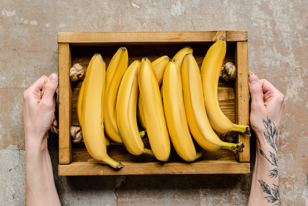 vue recadrée de la femme tenant des bananes mûres dans une boîte en bois sur une surface altérée - Photo, image