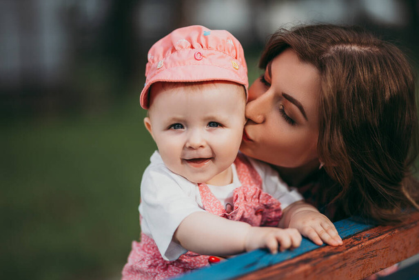ピンクの服を着た公園のベンチに座っている小さな子供を持つ若い美しい母親. - 写真・画像