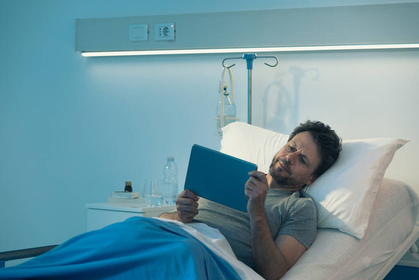 Patient insomniaque adulte allongé dans un lit d'hôpital la nuit, il regarde des films avec sa table numérique - Photo, image
