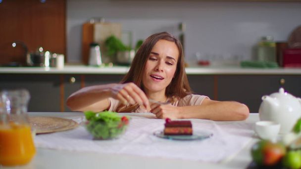 Mujer feliz comiendo pastel en lugar de ensalada en la cocina. Señora disfrutando de pastel en la mesa
 - Foto, Imagen