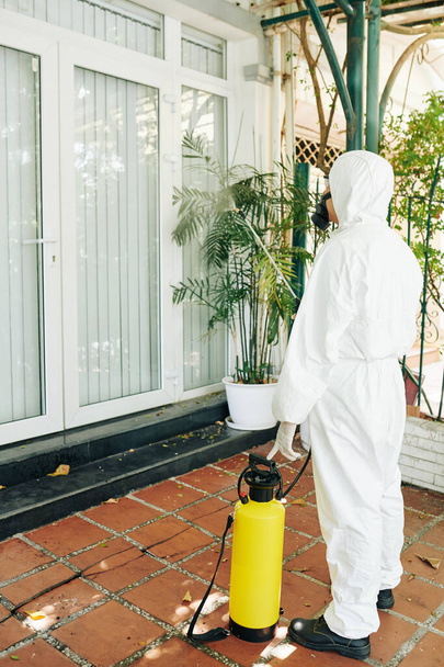 technicus in hazmat pak spuiten chemicaliën op muren en ramen van huis om virusverspreiding te voorkomen - Foto, afbeelding