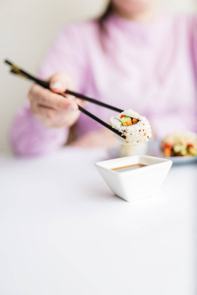Female dipping tasty sushi roll with chopsticks into bowl with soy sauce at the table. - 写真・画像