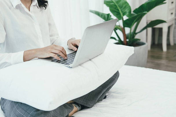 アジアの女性は寝室のベッドでコンピュータのノートパソコンを使ってる。家でリモートで働いてる。余暇生活 - 写真・画像