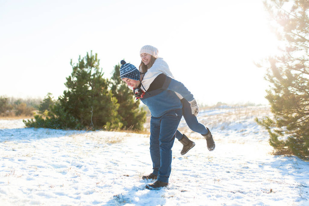 Loving couple play in the winter in the forest. Girl rides a guy in the background of the Christmas tree. Laugh and have a good time. - Photo, Image
