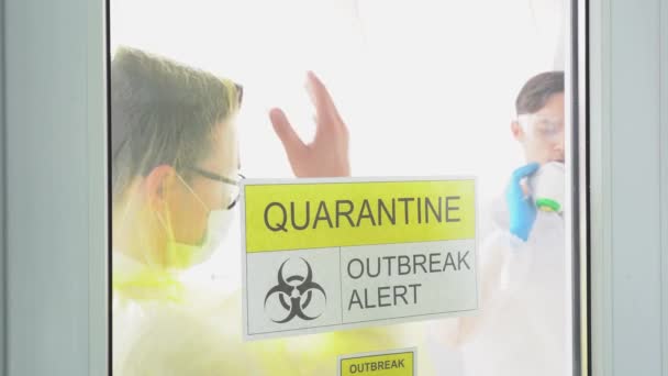 Two young guys in protective suits wearing medical face masks, glasses on background of white clinic ward. Men standing behind transparent door with prohibition sign: Quarantine, outbreak alert. - Footage, Video