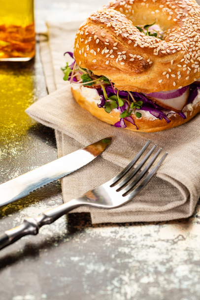 fresh delicious bagel with meat, red onion, cream cheese and sprouts on napkin with cutlery on textured surface - Foto, Bild