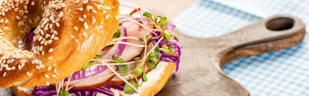 close up view of fresh delicious bagel with meat, red onion, cream cheese and sprouts on wooden cutting board near napkin, panoramic shot - Φωτογραφία, εικόνα