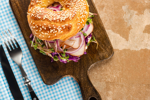 top view of fresh delicious bagel with meat, red onion and sprouts on wooden cutting board and plaid blue napkin with cutlery - Foto, Imagem