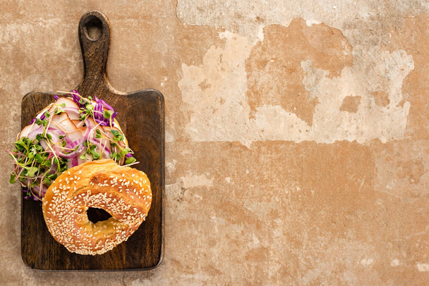 top view of fresh delicious bagel with meat, red onion and sprouts on wooden cutting board on aged beige surface - Foto, afbeelding