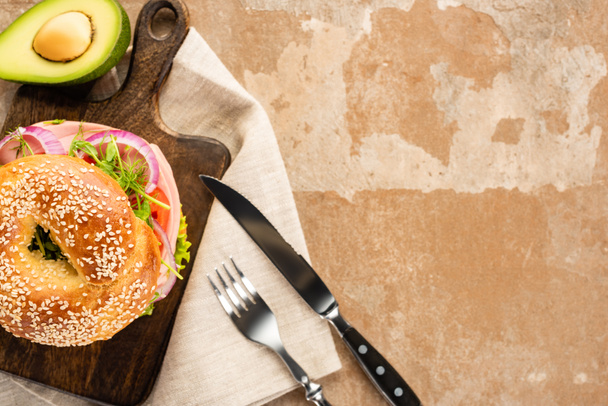top view of fresh delicious bagel on wooden cutting board on aged beige surface with avocado, cutlery and napkin - Photo, Image