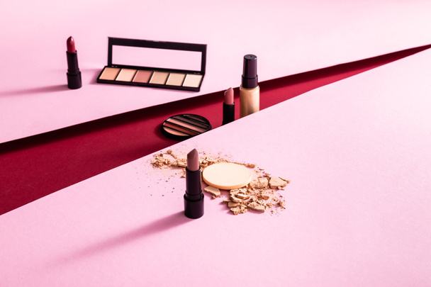 eye shadow palettes, lipsticks and cracked face powder near face foundation on pink and crimson  - Fotografie, Obrázek