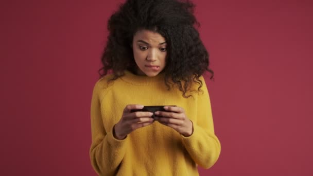 Young african concentrated positive woman isolated over dark red burgundy background play games by mobile phone make winner gesture - Záběry, video