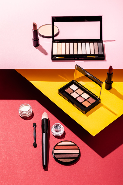 paletas de sombras de ojos y pinceles cosméticos cerca de lápices labiales y polvo facial sobre carmesí, rosa y amarillo
 - Foto, imagen