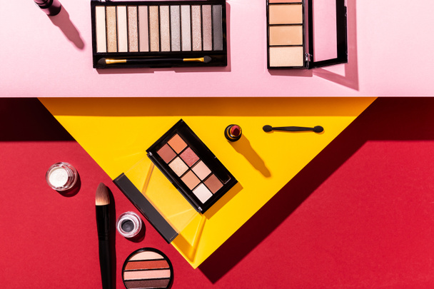 top view of eye shadow palettes and cosmetic brushes near lipstick on crimson, pink and yellow - Foto, afbeelding
