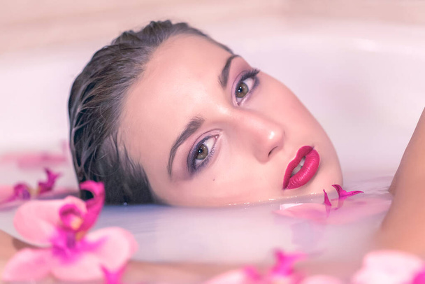gyönyörű lány pózol egy fürdő tele tejjel és virágokkal fotók - Fotó, kép