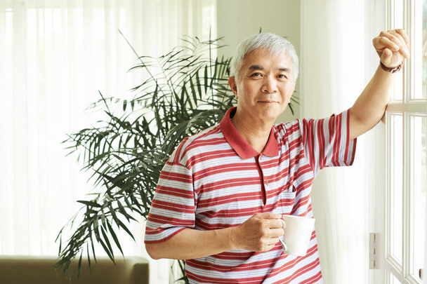 Boldog érett ázsiai férfi áll az ablakban, és iszik reggeli kávét otthon - Fotó, kép