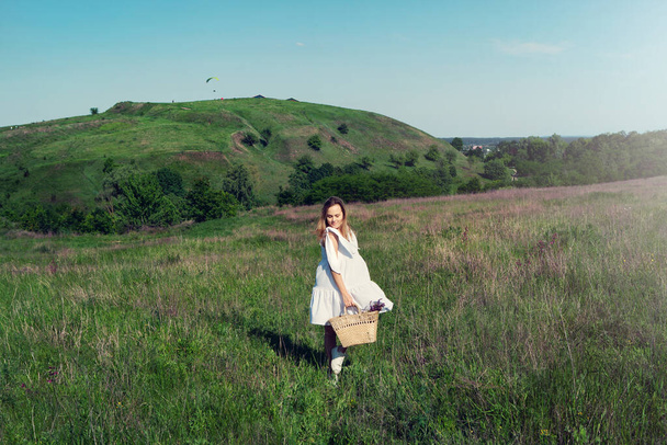 Hermosa chica en vestido blanco con bolsa de paja y flores silvestres corre en el prado
. - Foto, Imagen
