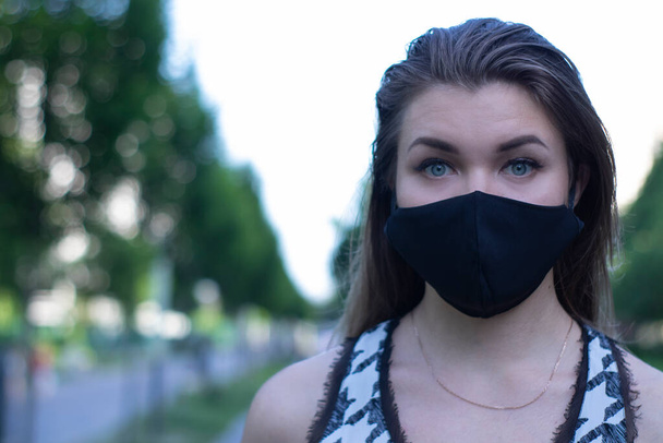 Pretty young blonde woman in medical black face mask. Wearing a t-shirt and jeans shorts. in a park. modern reality. covid-19 concept. copy space. - Photo, Image